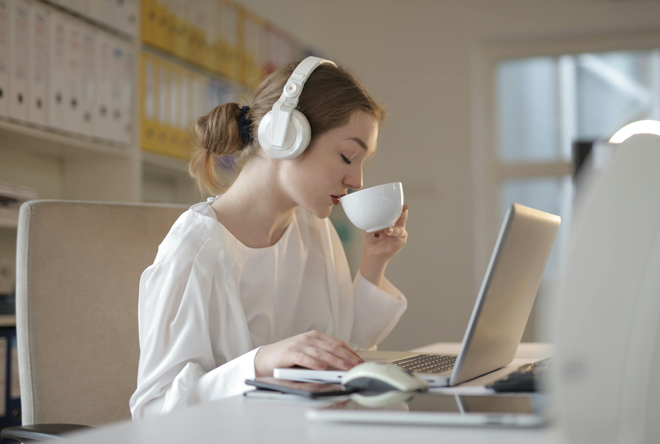 Woman wearing headphones and sipping coffee while working on a laptop at a desk.